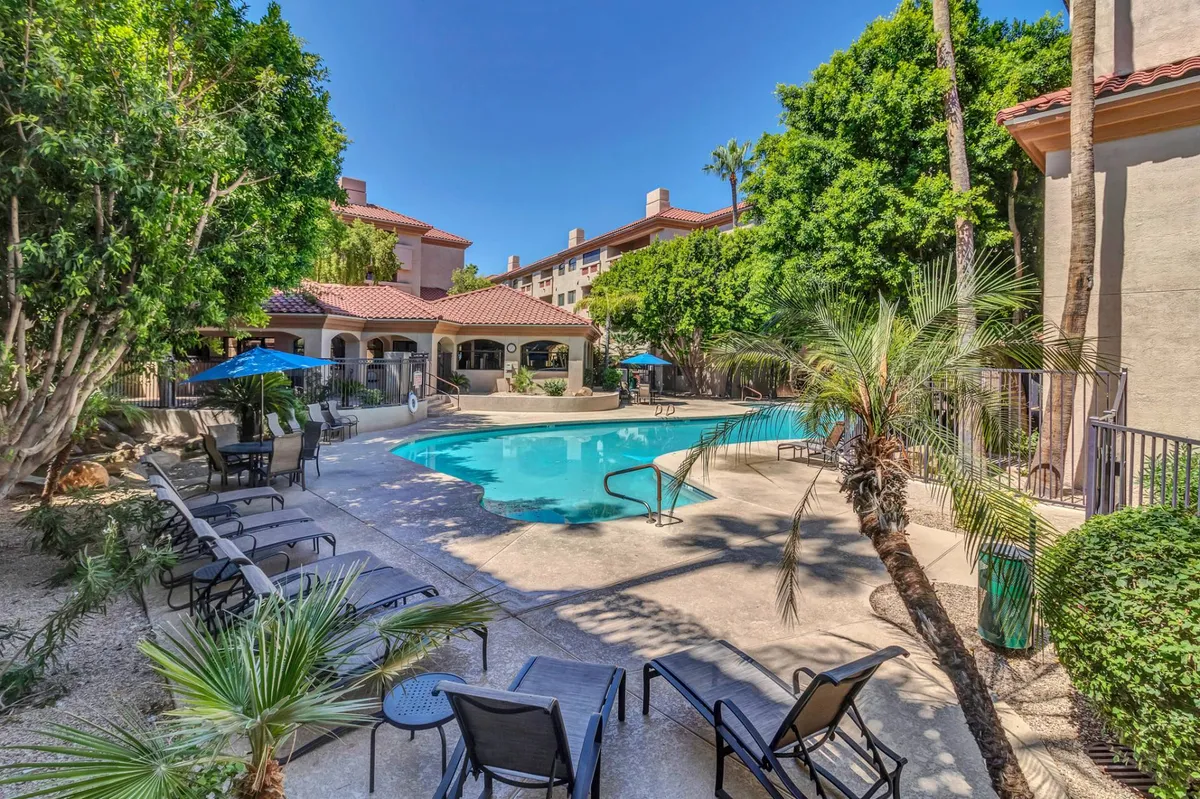 A sun-drenched outdoor swimming pool surrounded by palm trees and lounge chairs, with the resort's building architecture visible in the background. The tranquil setting invites guests for swims, sunbathing, and relaxing poolside, capturing the essence of a resort-style amenity.