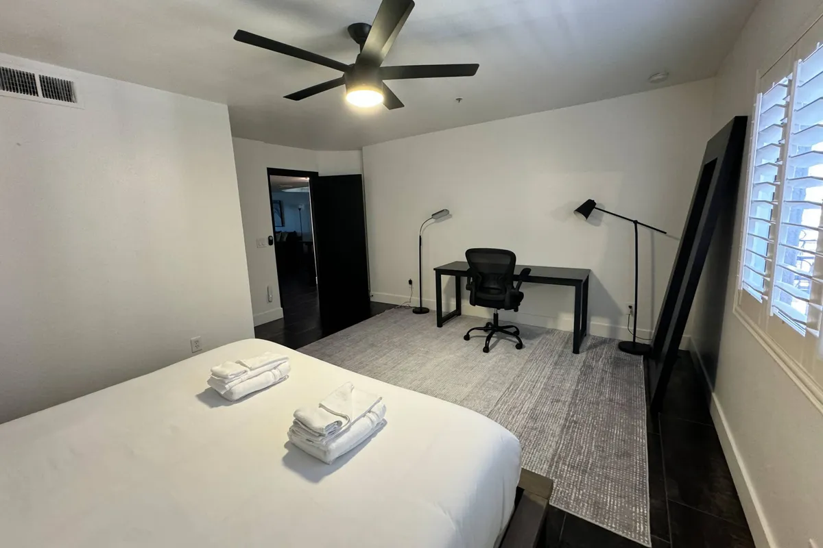 A modern bedroom setup with a bed, neatly folded towels, a desk and chair positioned for work or leisure, and a warm floor lamp. The scene feels contemporary and organized, providing a glimpse of a versatile space perfect for longer stays.