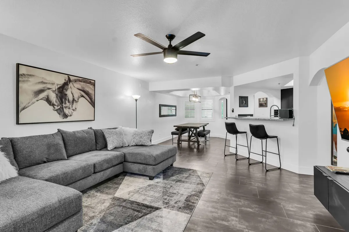 This image captures a spacious open-plan living area with a large sectional sofa at the center, framed artwork of a horse on the wall, a dining table, and bar stools at the kitchen counter. The scene radiates warmth and comfort, perfect for gatherings, family relaxation, or casual dining and entertainment.