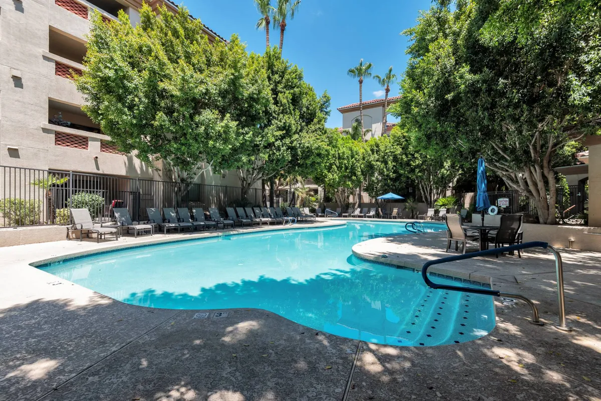 A sunlit community swimming pool surrounded by sleek lounge chairs and mature, leafy trees creates a tranquil oasis for relaxation and recreation. The area is meticulously maintained, inviting guests to swim, sunbathe, or socialize in comfort.