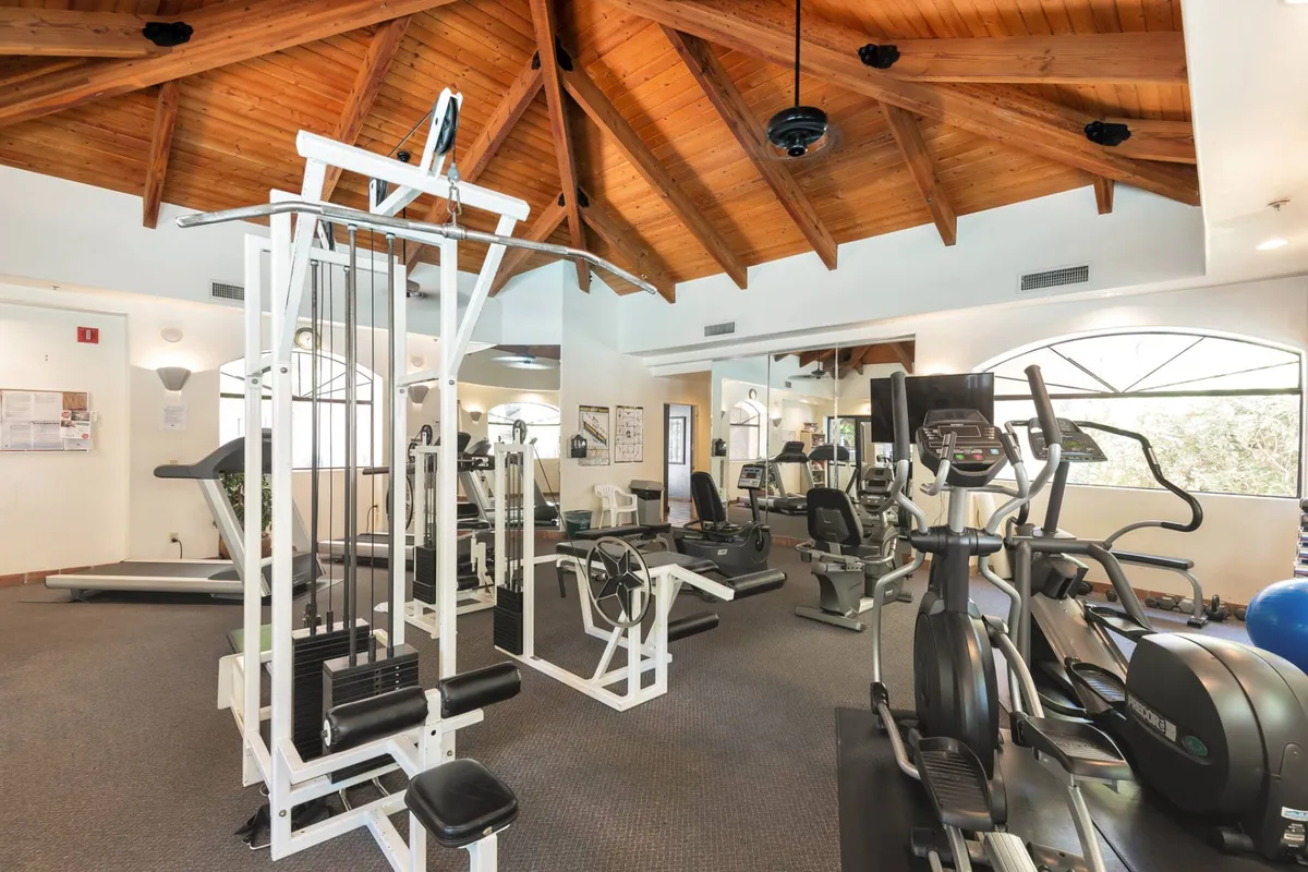 A bright, modern fitness center equipped with a weightlifting machine, elliptical trainer, treadmill, and a mirrored wall. The space is designed for group classes, personal training, and individual workouts, promoting an active lifestyle.