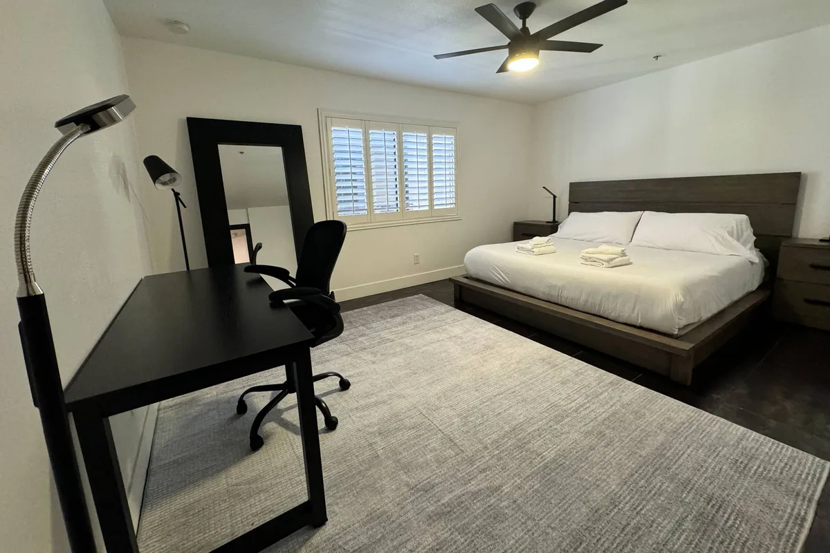 A tranquil primary bedroom with a king-size bed, a dedicated desk and chair for work or journaling, a bedside lamp, and a mirror—all set in a serene, softly lit environment ideal for relaxation or productivity. The space looks thoughtfully organized with plush linens and ample storage.