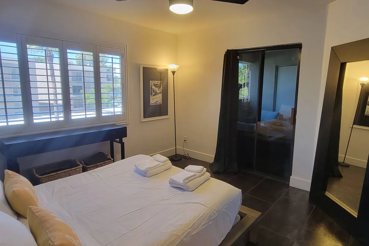 A tranquil bedroom scene featuring a neatly made bed with crisp towels placed atop, a soft floor lamp casting inviting light, decorative wall art, and a framed mirror. Ample natural light streams in, suggesting a restful space with a hint of morning sunlight.
