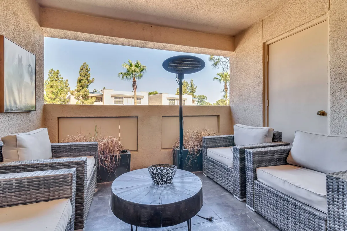 A tranquil private balcony scene featuring wicker chairs, a circular table, decorative plants, and an outdoor heater. The setting is perfect for relaxing with morning coffee or evening drinks while enjoying fresh air and views.