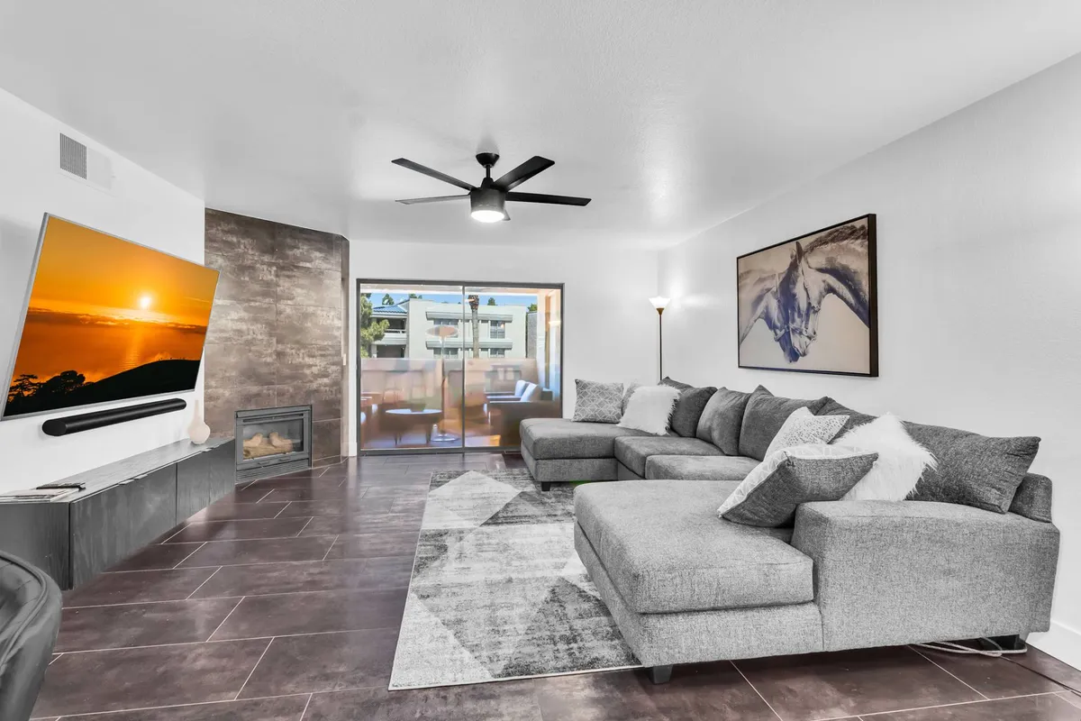 The image showcases a stylish, contemporary living room with a spacious sectional sofa facing a mounted flat-screen TV. Modern artwork decorates the walls, and natural light pours in through a sliding glass door that opens onto a private balcony, hinting at sunset views. The space feels inviting, perfect for relaxing, socializing, or enjoying a cozy evening indoors.