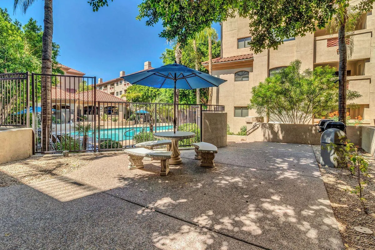 A communal BBQ area with an outdoor table shaded by an umbrella, pool views in the background, and lush landscaped greenery. The space is set up for outdoor meals, socializing, and enjoying the community environment.