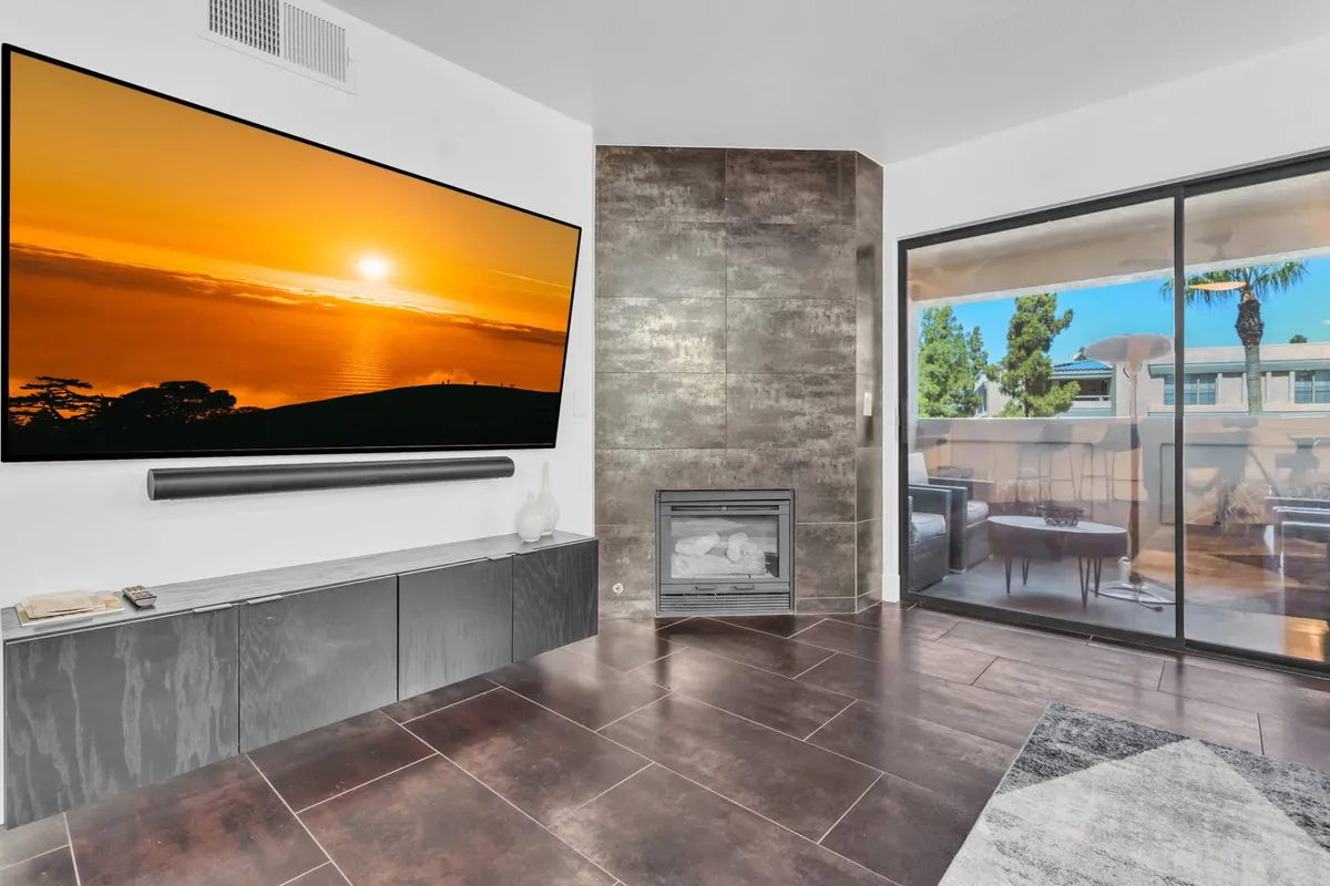 The image presents a stylish living room with a large flat-screen TV, a modern gas fireplace, and expansive glass doors opening to the balcony, blending indoor relaxation with access to outdoor space.