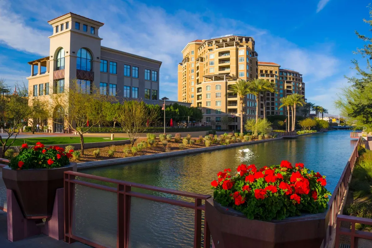 Modern buildings are framed by lush greenery, colorful flower planters, and a reflective water surface. The scene conveys a vibrant, welcoming urban atmosphere, ideal for business travelers who appreciate access to dining and social spaces within a sophisticated environment.